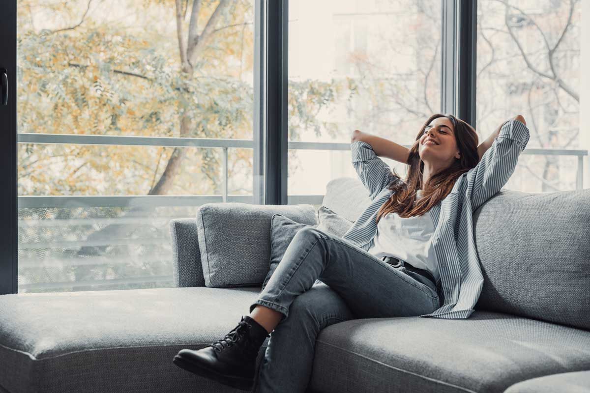Woman relaxing on a gray couch with arms behind her head near a large window; fall foliage visible.