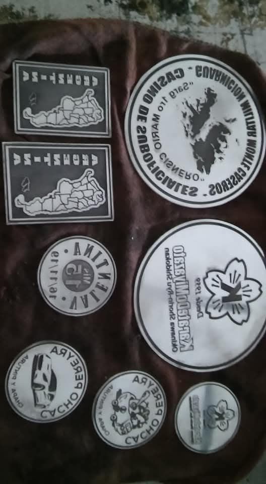 Varios logotipos circulares y rectangulares en blanco y negro, con forma de sello, dispuestos sobre un fondo de tela oscura.