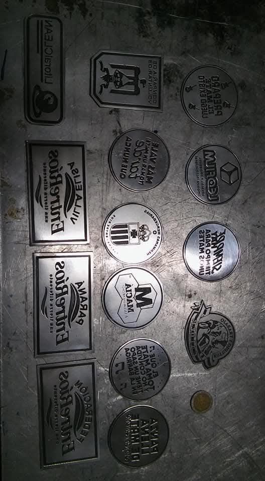 Letrero metálico con logotipos circulares y rectangulares dispuestos sobre una pared gris desgastada.