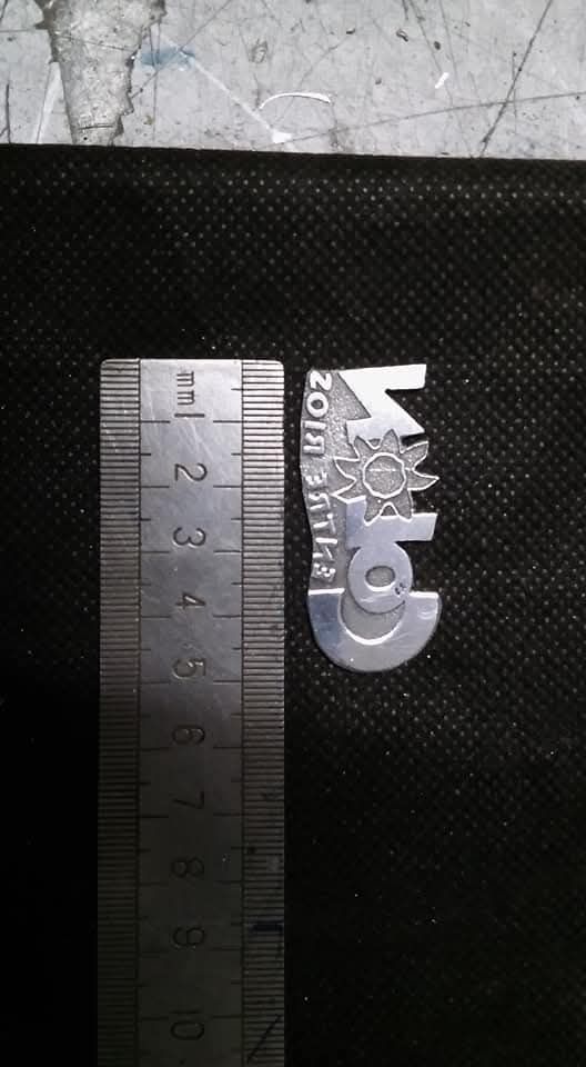 Pequeño logotipo de metal plateado junto a una regla sobre una alfombrilla negra texturizada, visto de cerca.