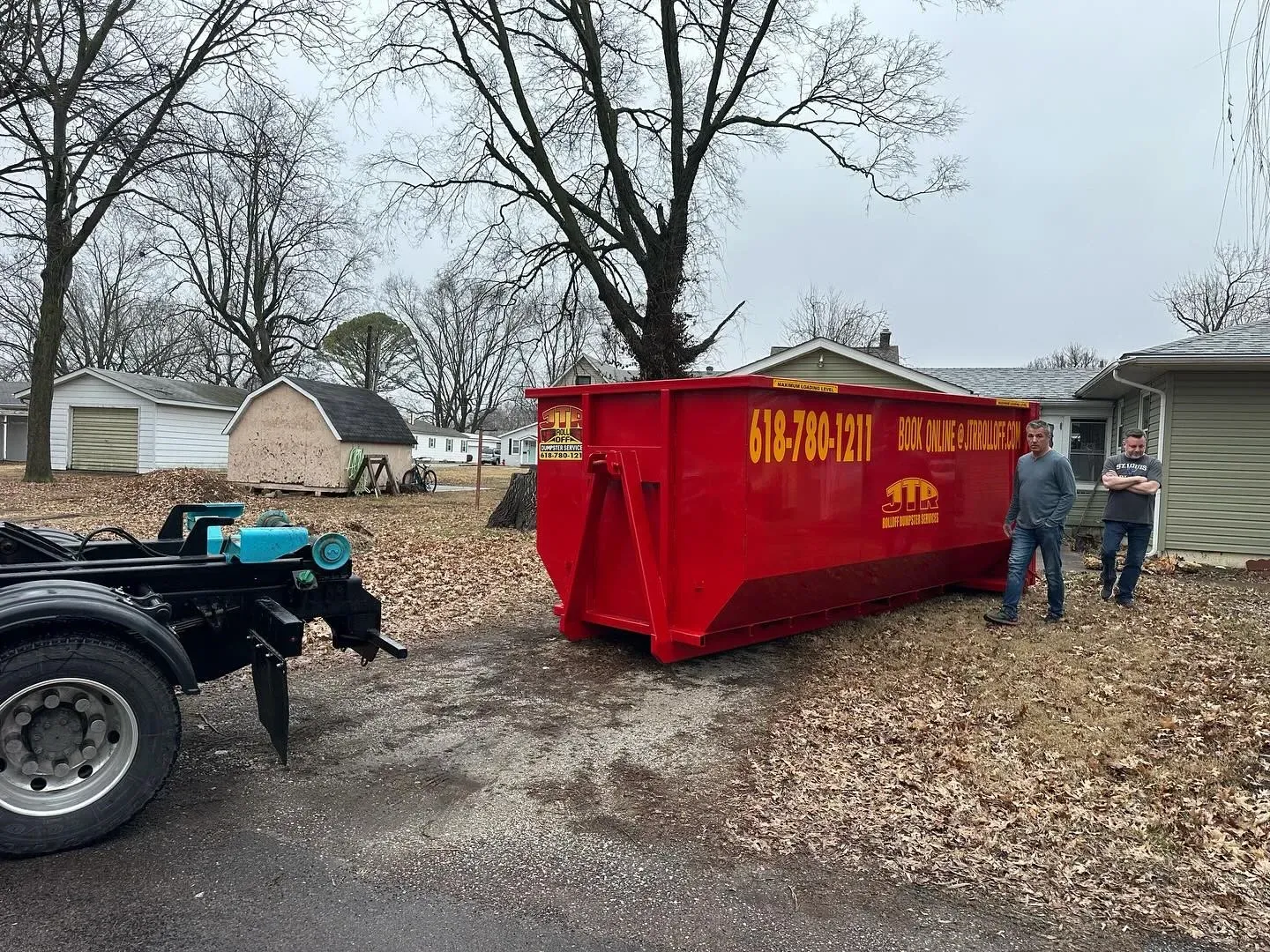 A red dumpster is sitting on the side of the road next to a trailer.