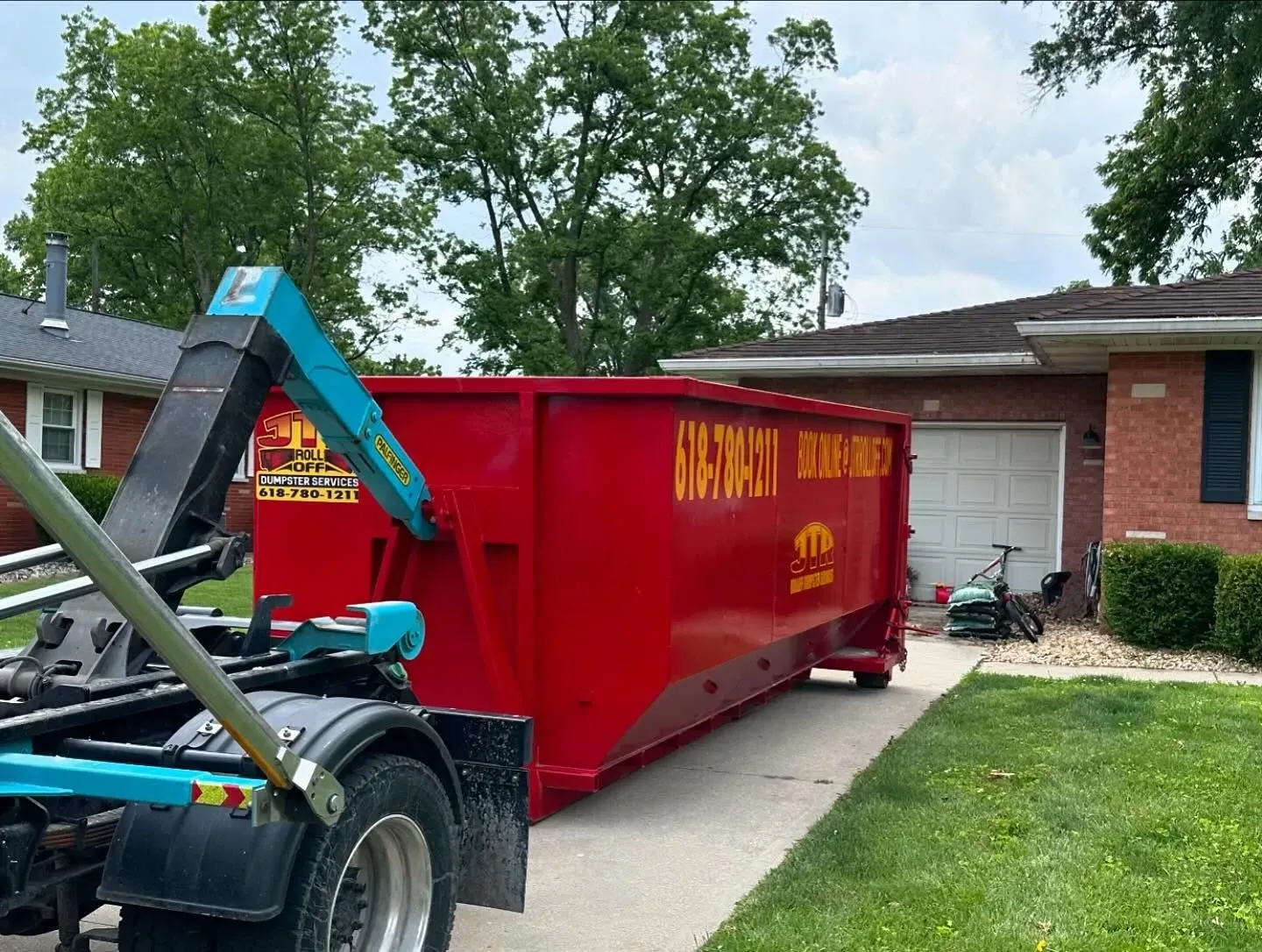 A red dumpster is being towed by a dumpster truck in front of a house.