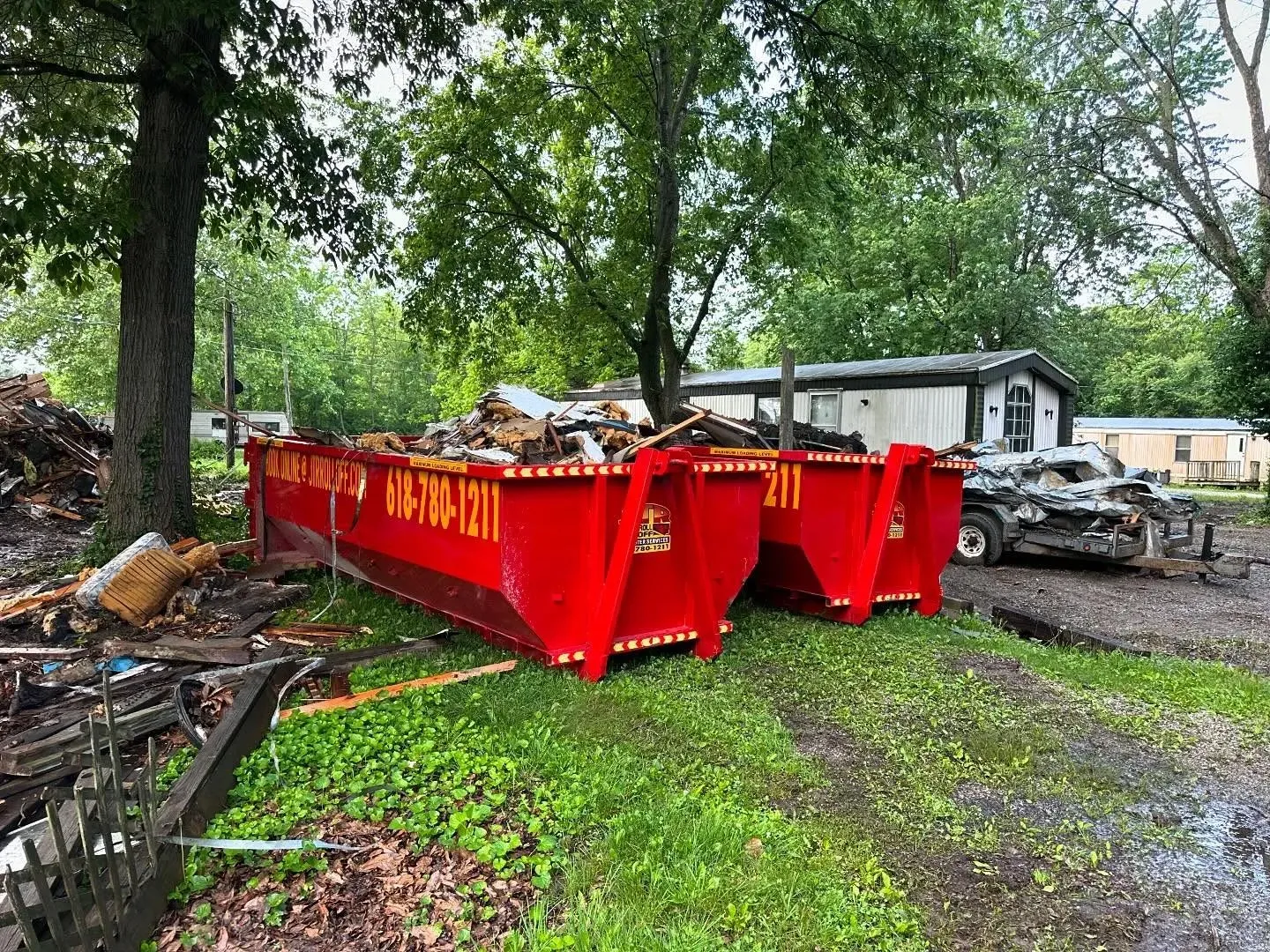 Two red dumpsters are sitting in the grass in front of a house.
