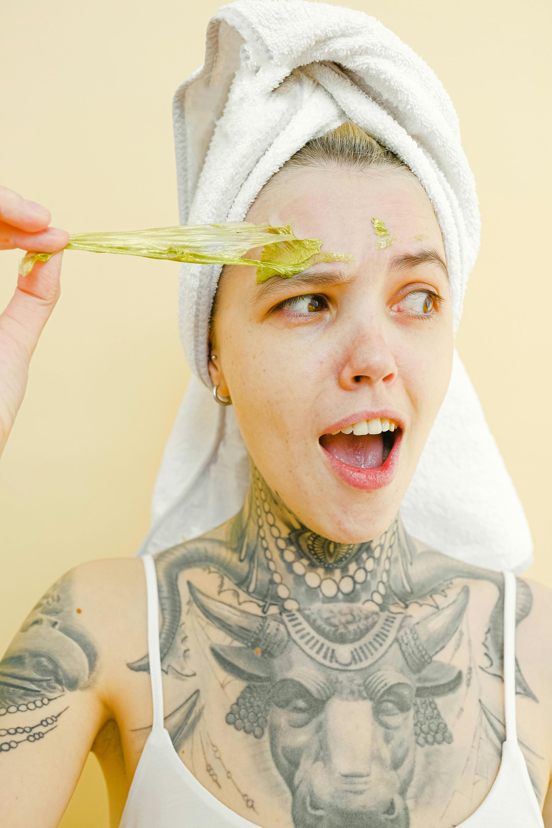 Woman with towel on head applying green facial mask, mouth open, showing chest tattoos.