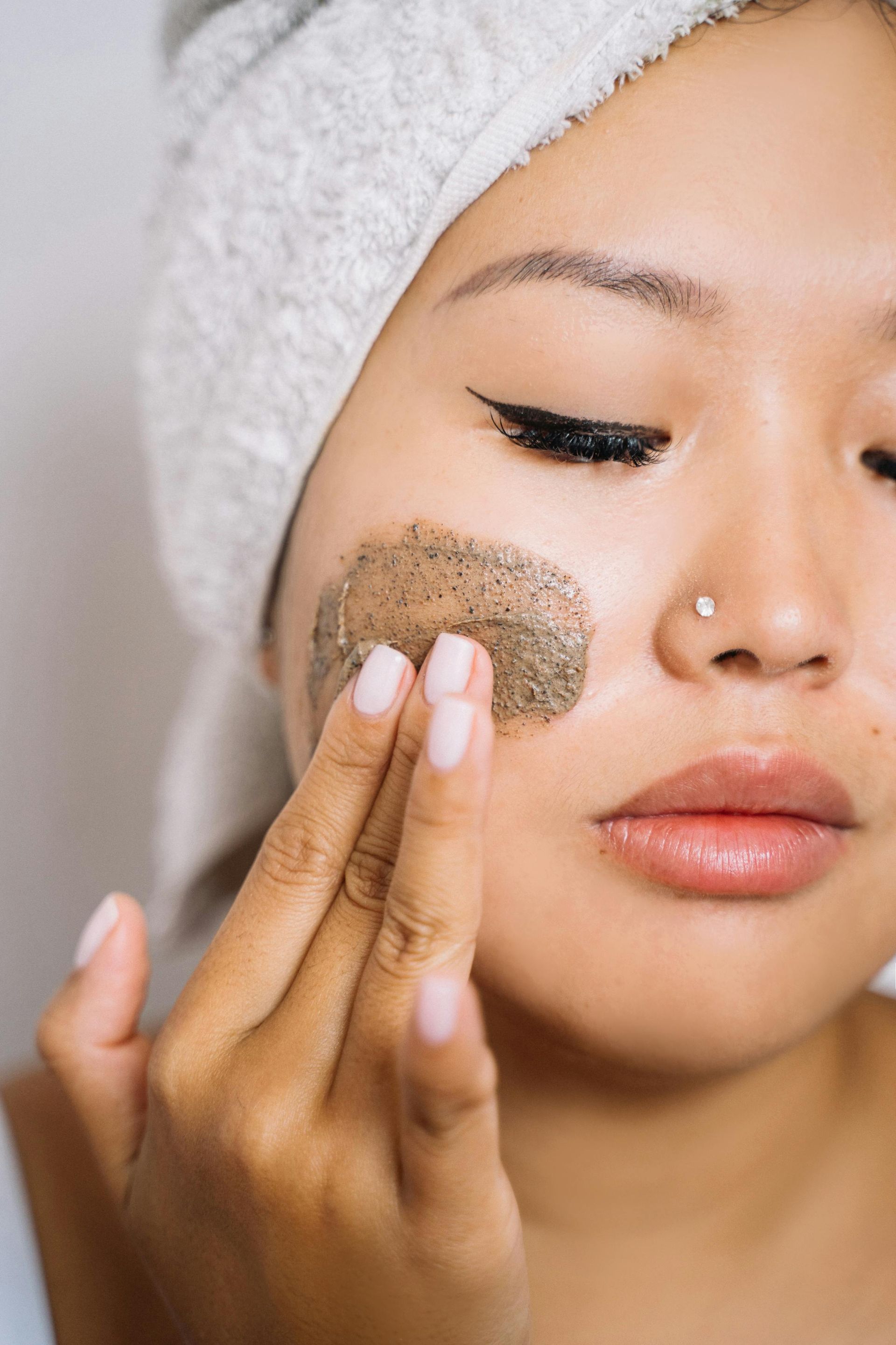 Woman applying a green facial scrub to her cheek with closed eyes and a towel wrapped around her head