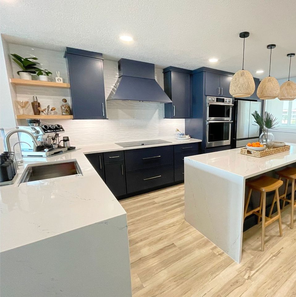 Navy blue kitchen with white countertops and island, wooden floors, and woven pendant lights.