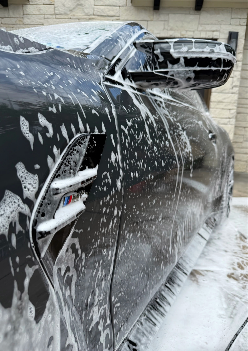 Black car covered in soap suds during a wash, parked beside a building and snow-covered ground