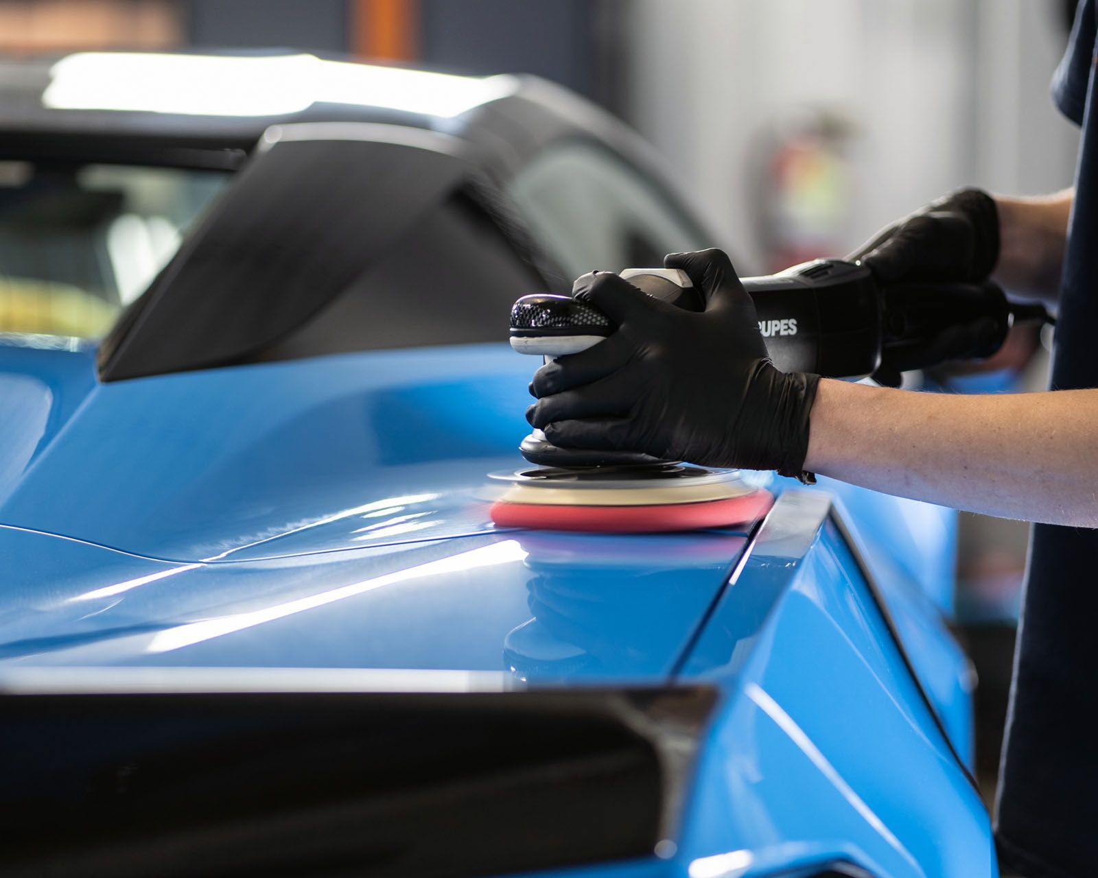 Gloved hands polishing a blue sports car with a buffer in a garage