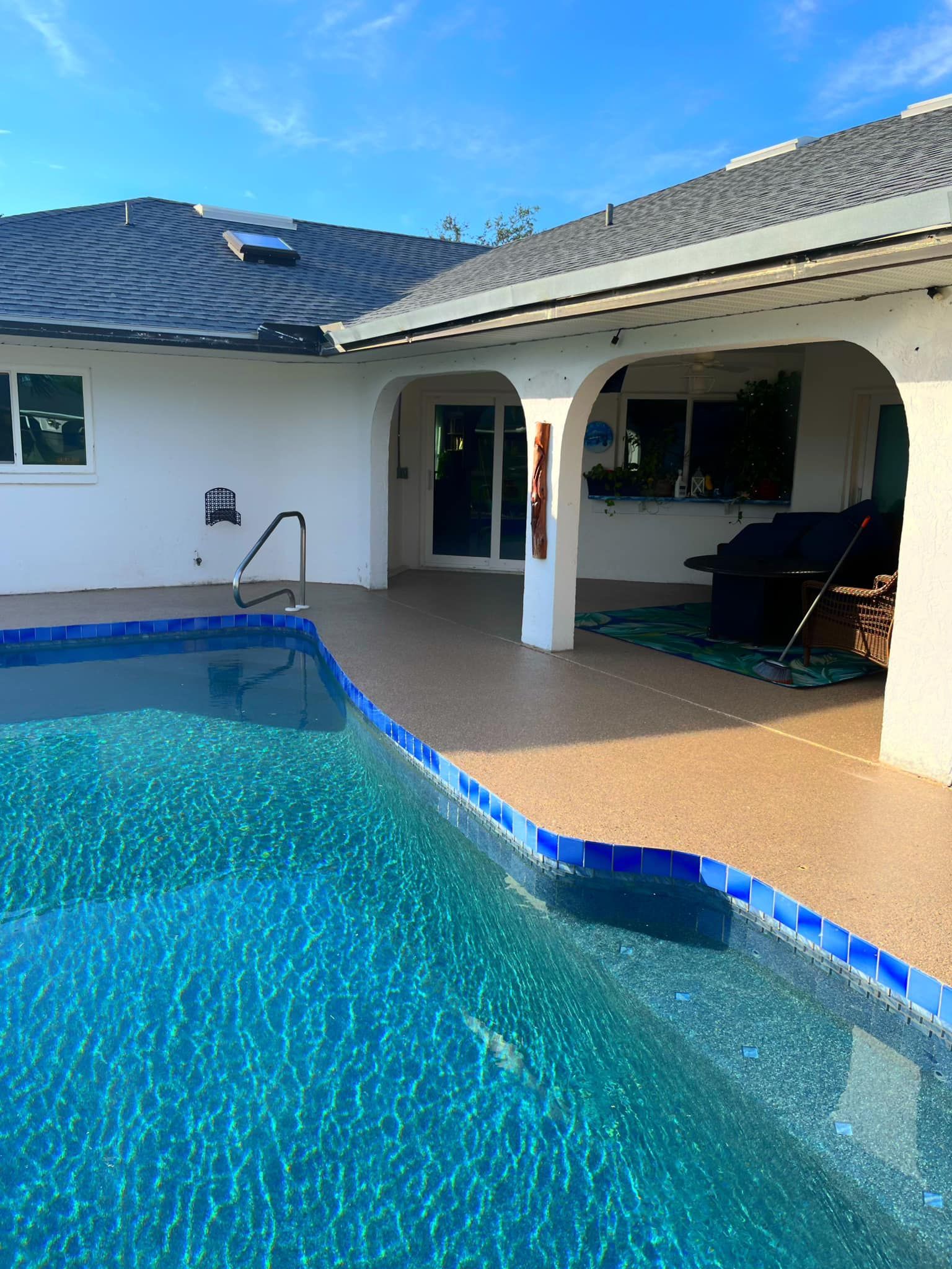 Pool with blue water next to a brown patio under a white house with a dark roof.