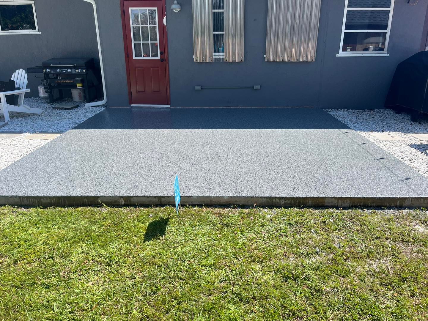 Concrete patio with dark grey surface, adjacent to grass, bordered by white stones, and a red door.