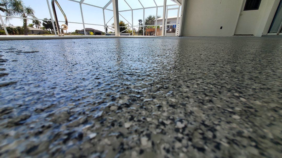 Low-angle view of a speckled, glossy concrete floor. The background includes a pool, enclosure, and a sunny sky.