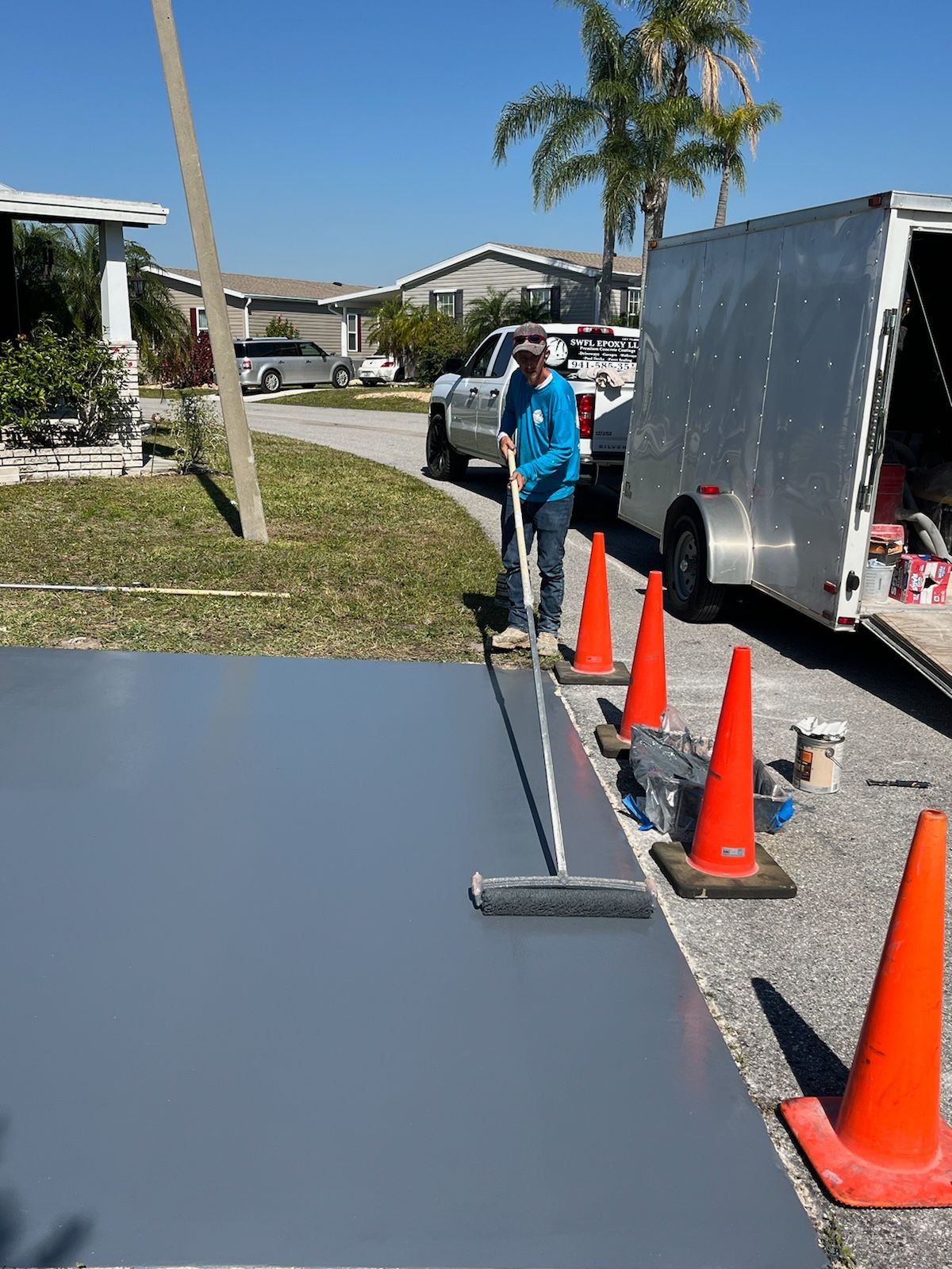 Person rolling gray paint onto a driveway, with orange cones, trailer, and houses in the background.