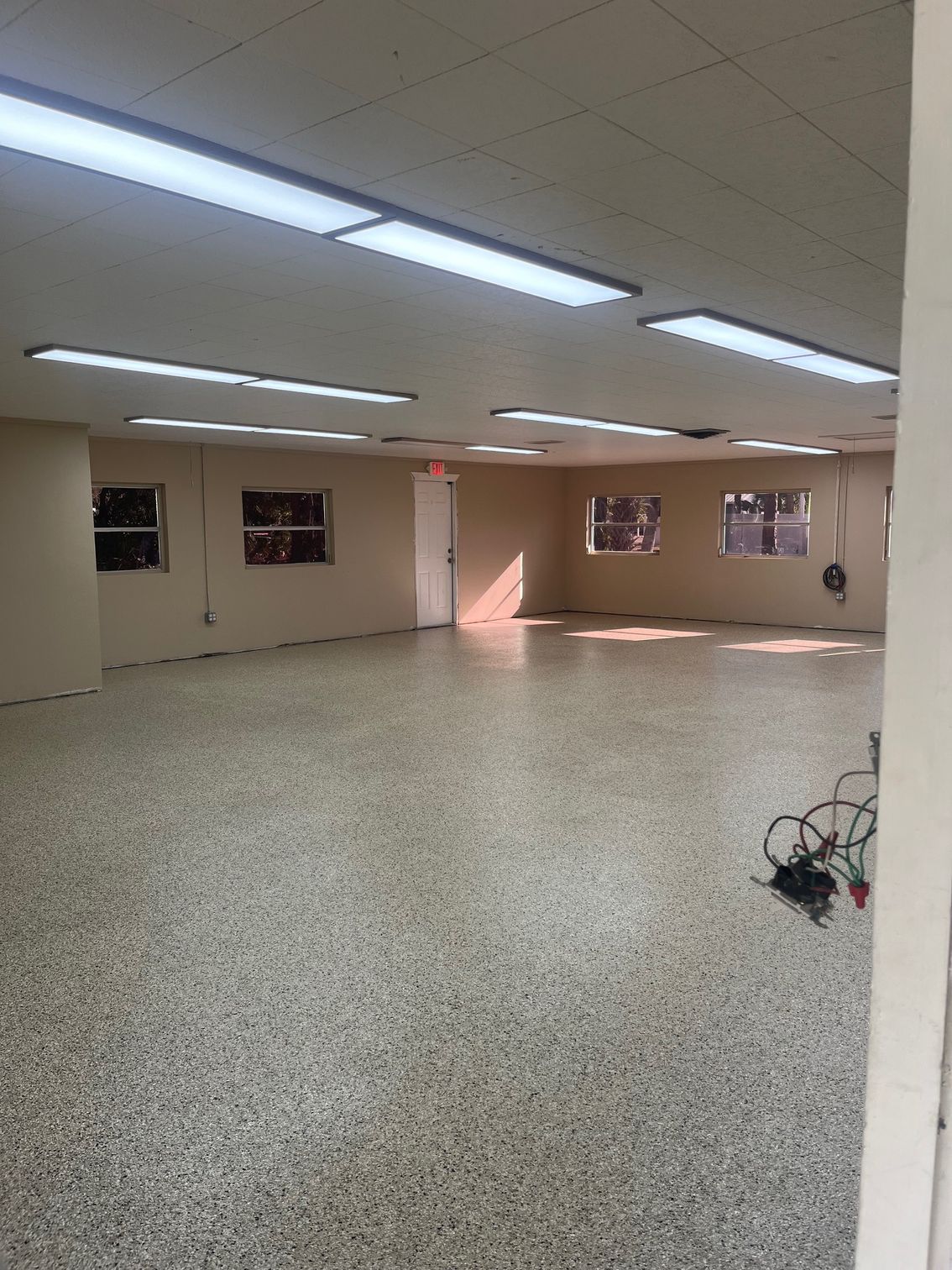 Empty room with speckled floor, beige walls, and fluorescent lights. Door and windows visible.