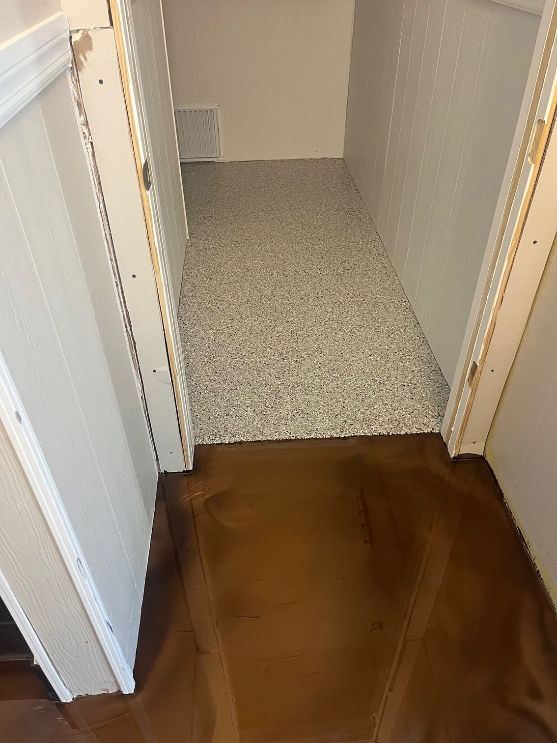 Brown floor leads to a hallway with speckled flooring, framed by white doorways.