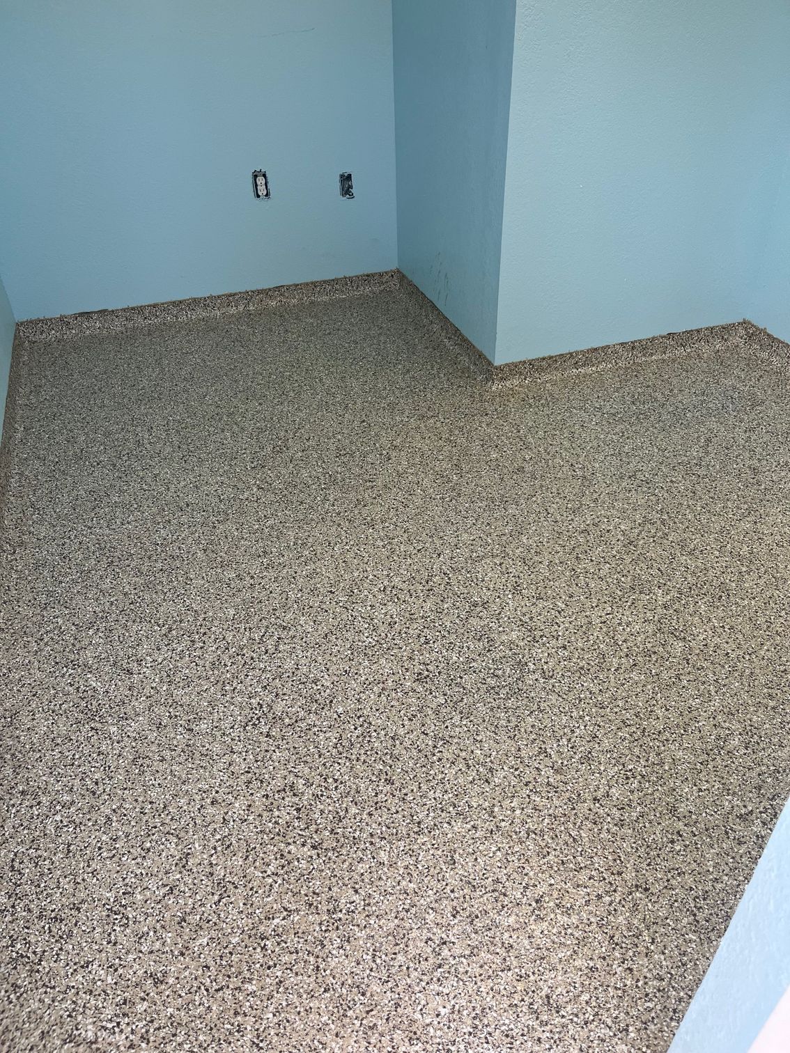 Corner of room with blue walls and speckled epoxy floor. Two electrical outlets are visible.