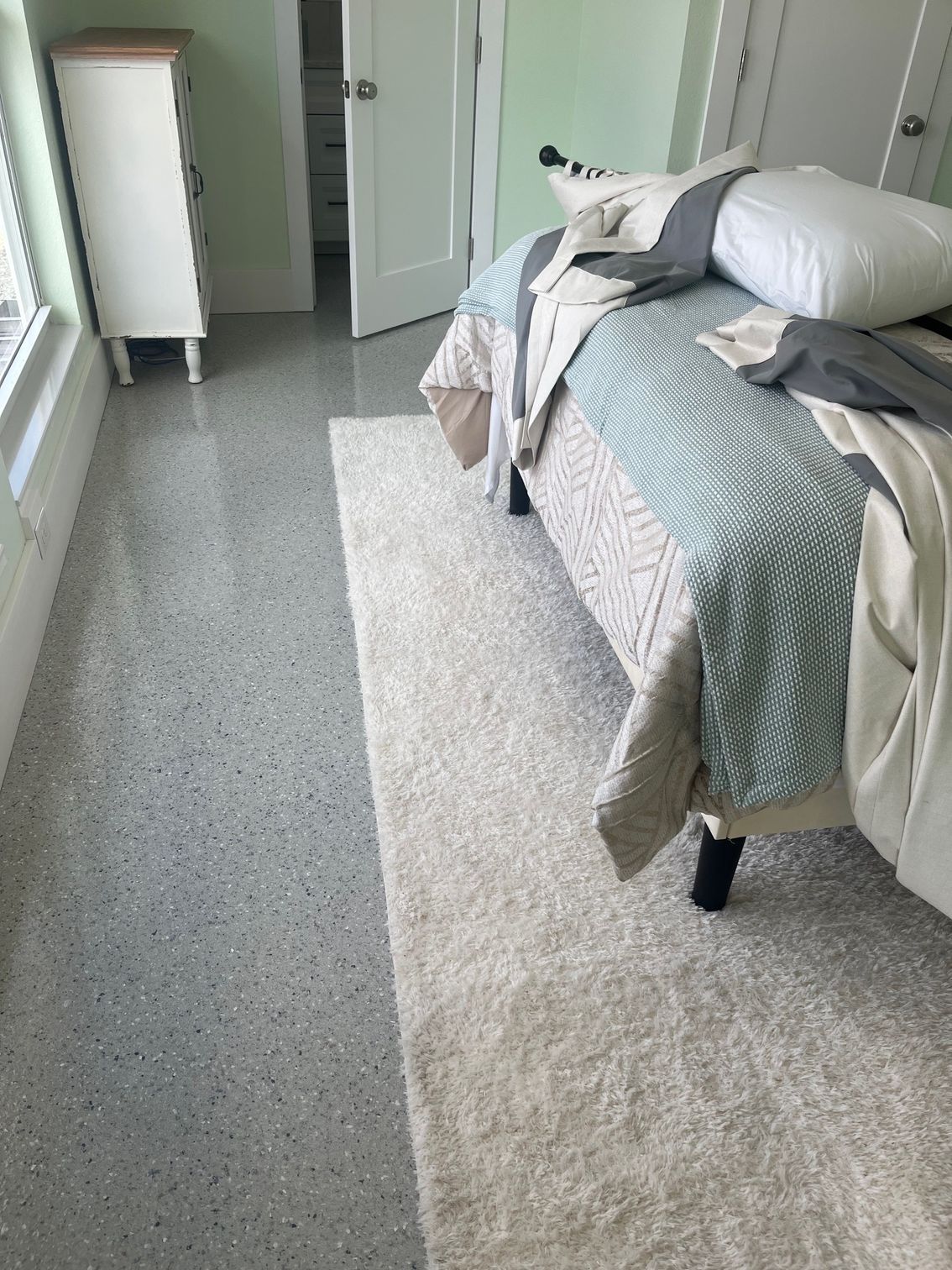 Bedroom with terrazzo floor, white rug beside bed with blue and beige bedding, white dresser.