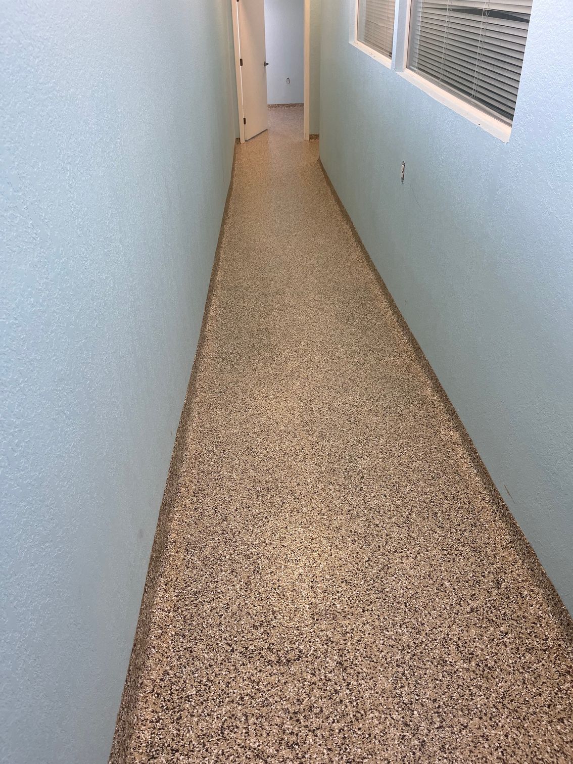 Narrow hallway with light blue textured walls and speckled flooring. White trim around a window.