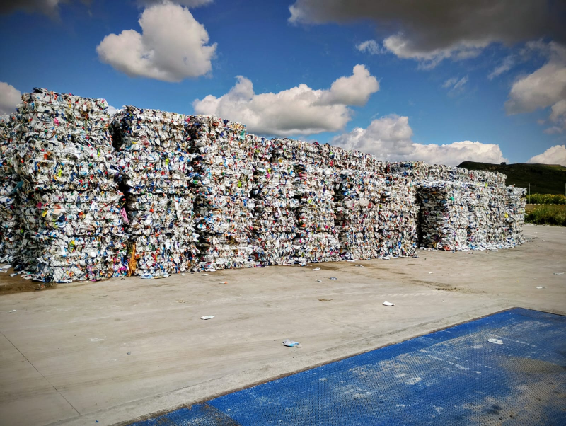 Sotto un cielo azzurro con nuvole bianche, pile di balle di rifiuti di plastica compattati giacciono su una superficie di cemento.