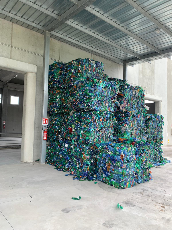 Grandi pile di bottiglie di plastica verde compresse, stoccate in un'area industriale esterna coperta.