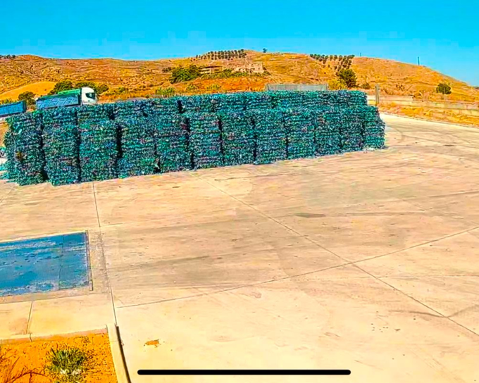 Una grande pila di bottiglie di plastica compresse, di colore verde, si erge su un piazzale di cemento, sullo sfondo di un paesaggio collinare e arido.