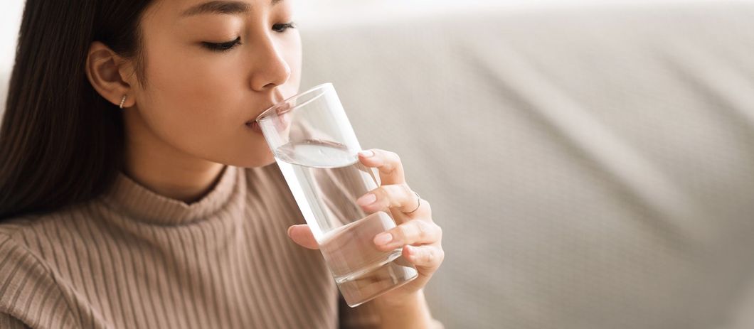 Una donna con un maglione beige beve acqua da un bicchiere trasparente seduta su un divano.