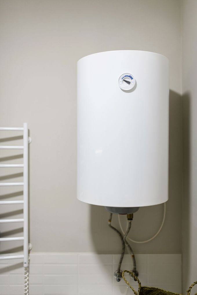 Scaldabagno bianco a parete con tubi, installabile in un bagno piastrellato o in un locale di servizio.