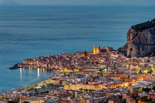 Città costiera al crepuscolo, con il centro storico illuminato in cima a una collina e il porto affacciato sul mare.