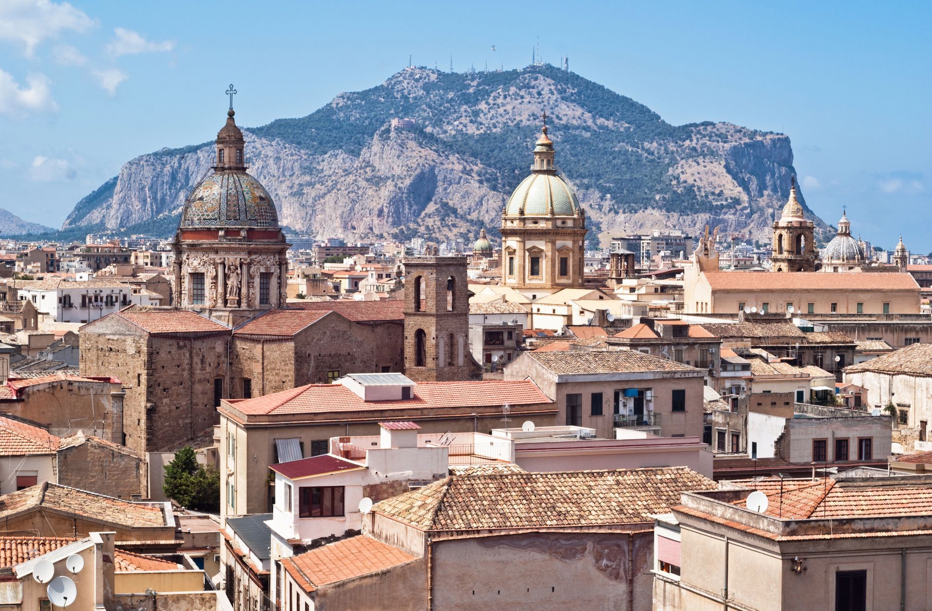 Panorama storico della città con tetti di terracotta, cupole di chiese e un'imponente montagna sullo sfondo.