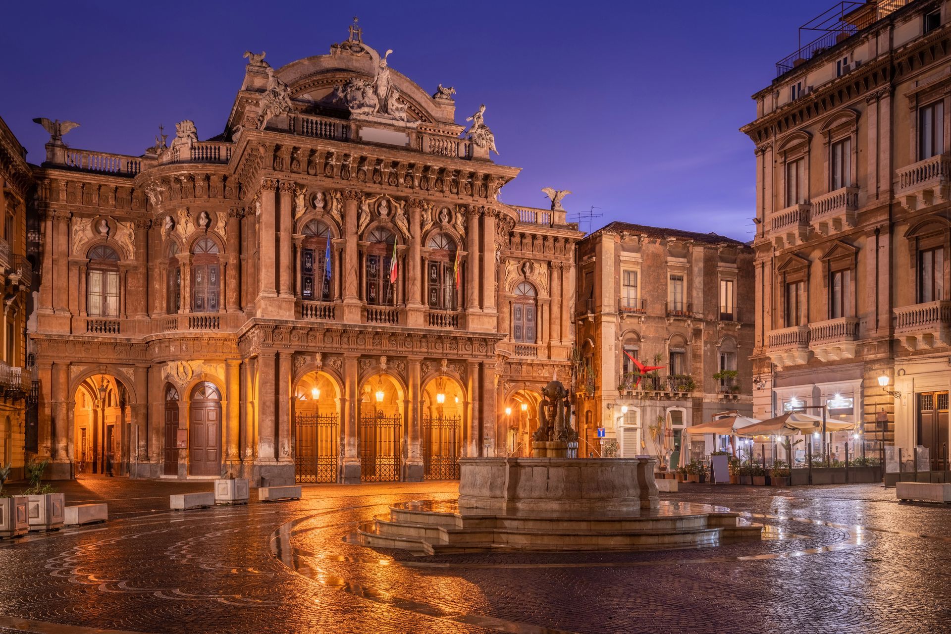 Teatro dell'opera storico illuminato e fontana in una piazza cittadina resa scivolosa dalla pioggia al crepuscolo