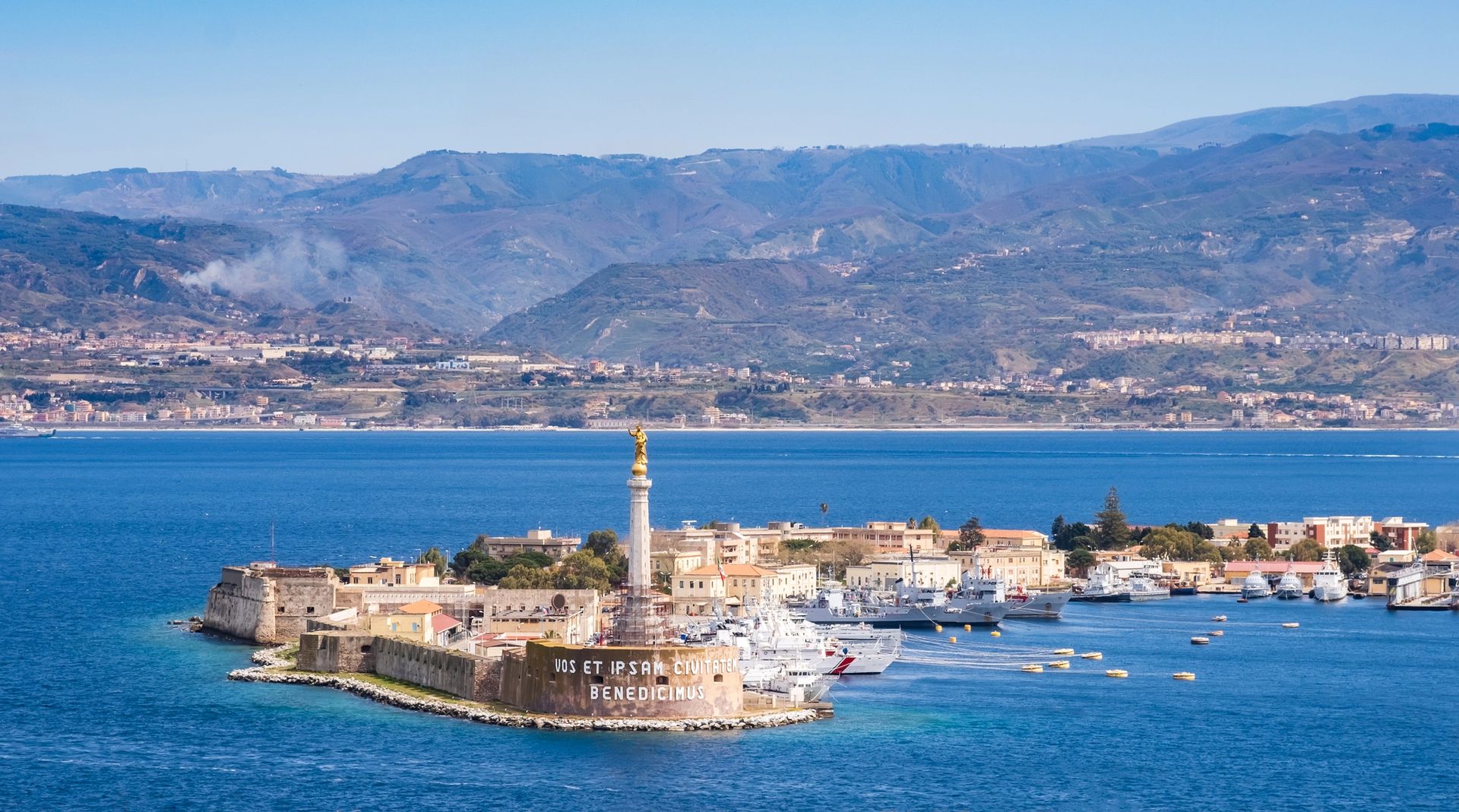 Fortezza costiera su un'isola rocciosa con un faro, mare blu e montagne sullo sfondo.