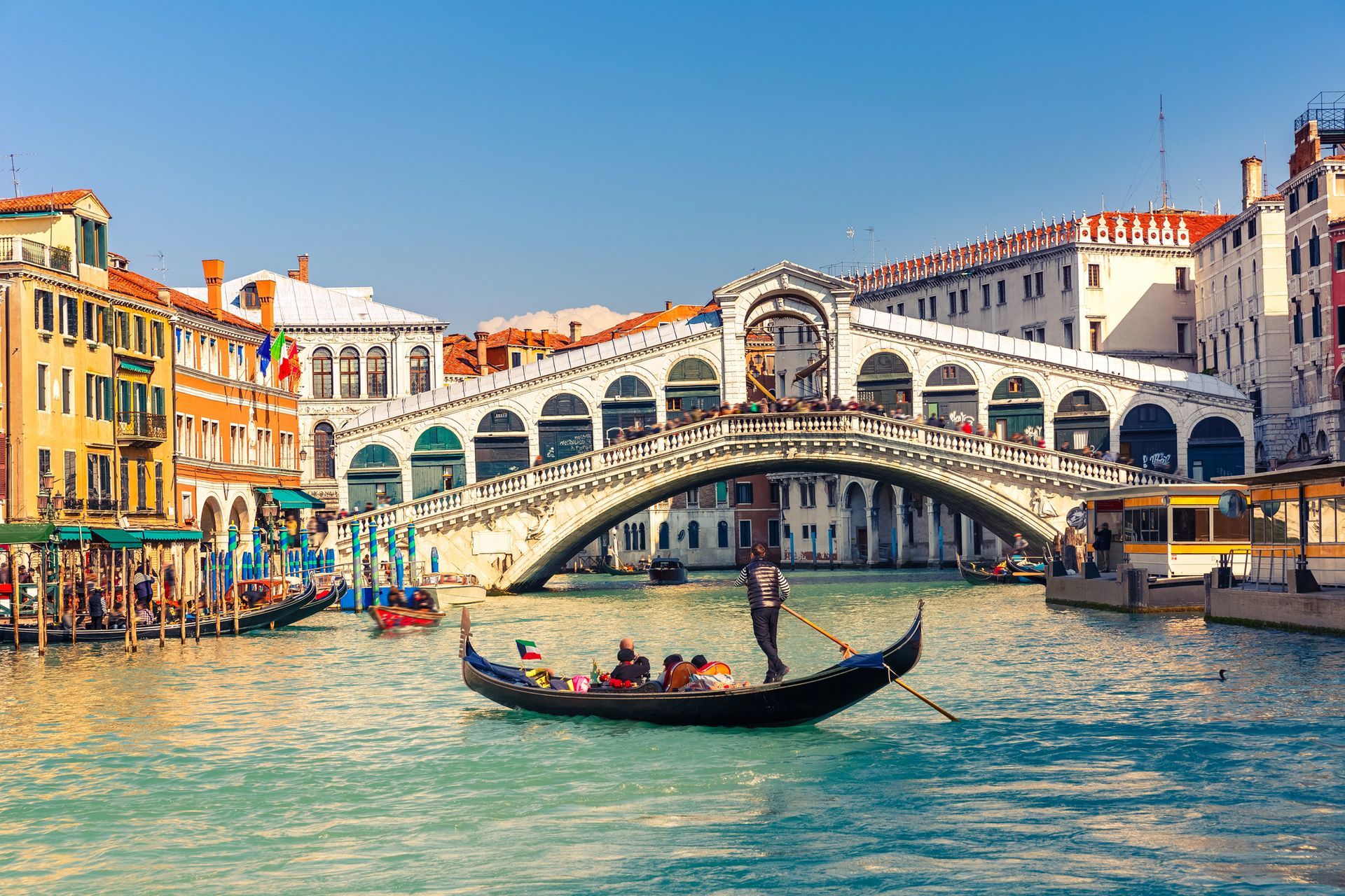 Il Ponte di Rialto di Venezia, che attraversa un canale con una gondola e palazzi colorati in una giornata di sole.