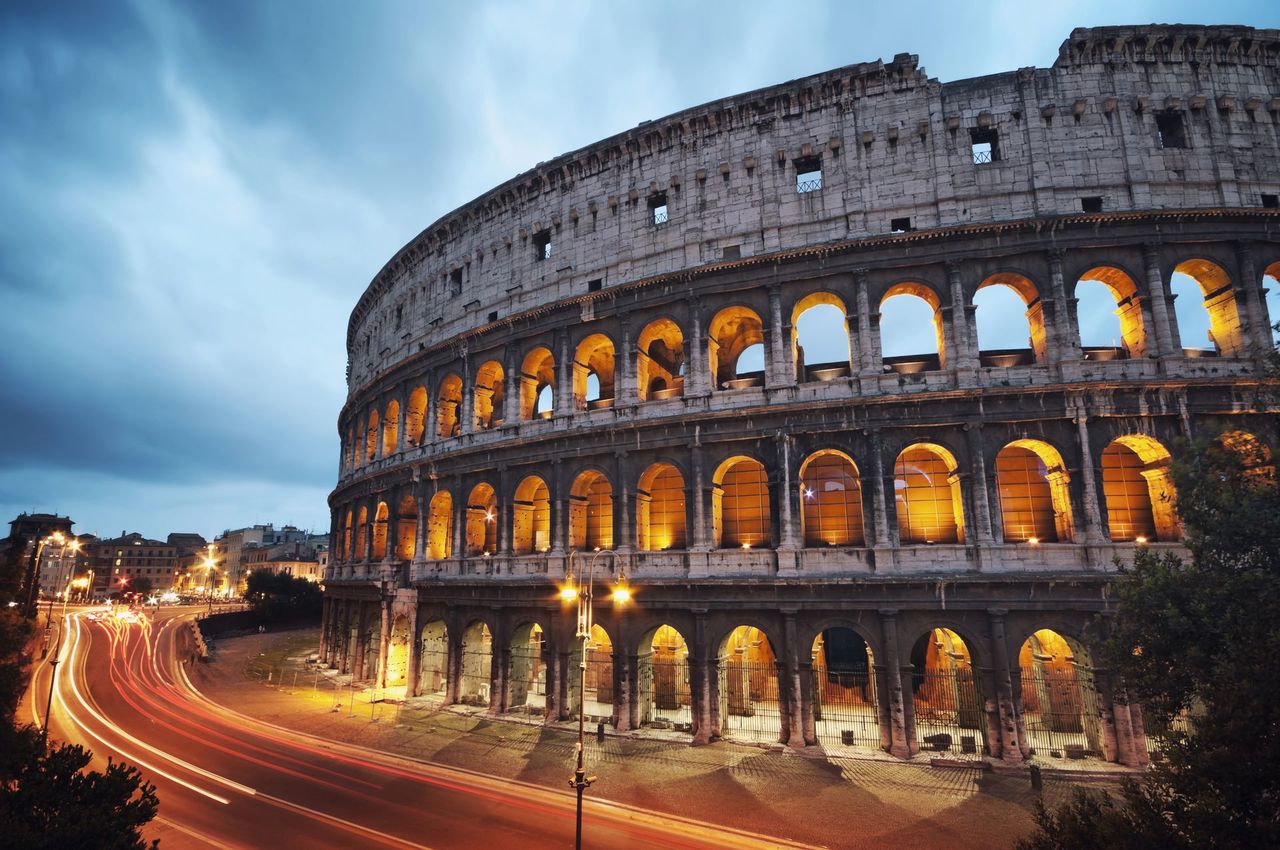 Colosseo illuminato al crepuscolo, con scie luminose lasciate dal traffico di passaggio nelle vicinanze.
