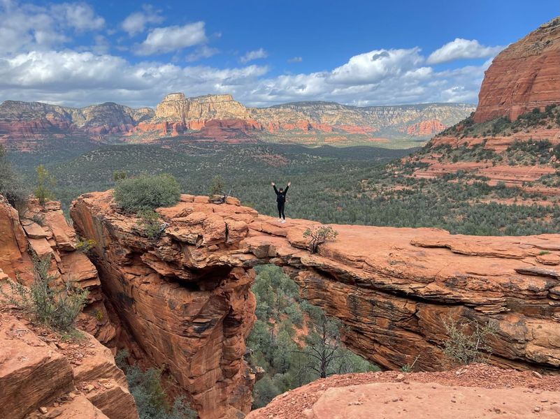 A person with arms raised stands on Devil's Bridge in Sedona, surrounded by red rock formations and a green valley.