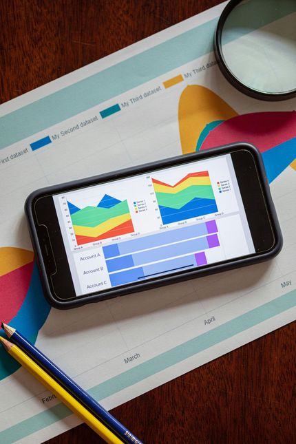 A smartphone displays colorful business charts on a desk with a magnifying glass and pencils on a paper graph background.