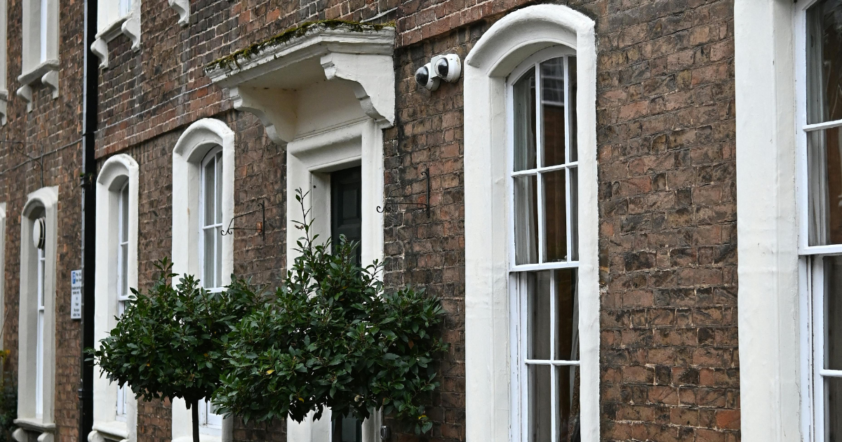 A red brick building facade with white-framed, arched windows, a doorway with a decorative canopy, and two potted bushes.