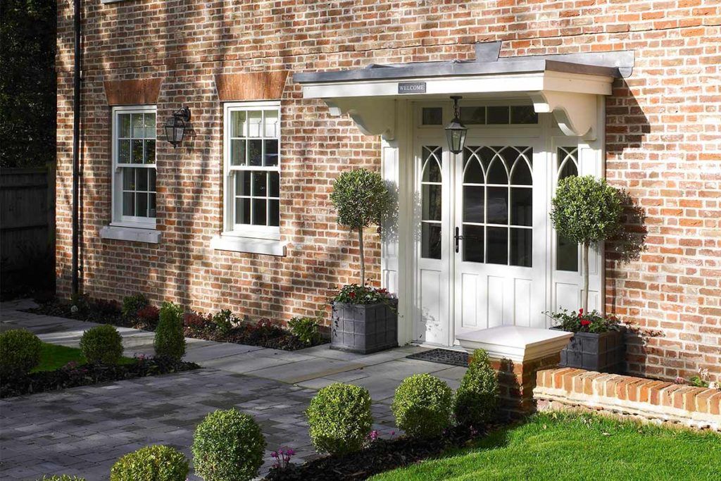 A brick home exterior with a white door featuring gothic-style glass panes, a canopy, and several manicured topiary trees.