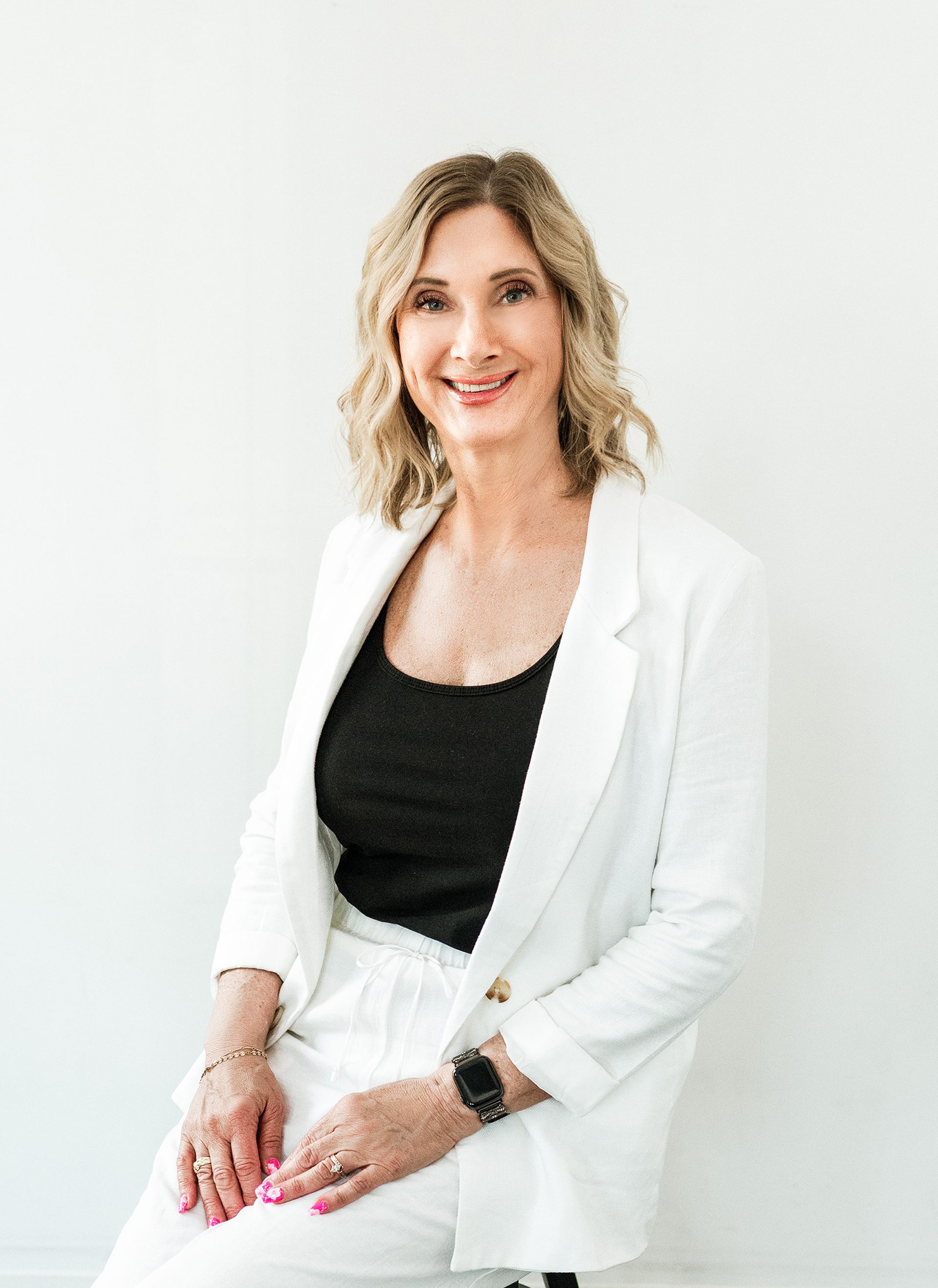 Woman in white suit sits on a stool, smiling, against a white wall.