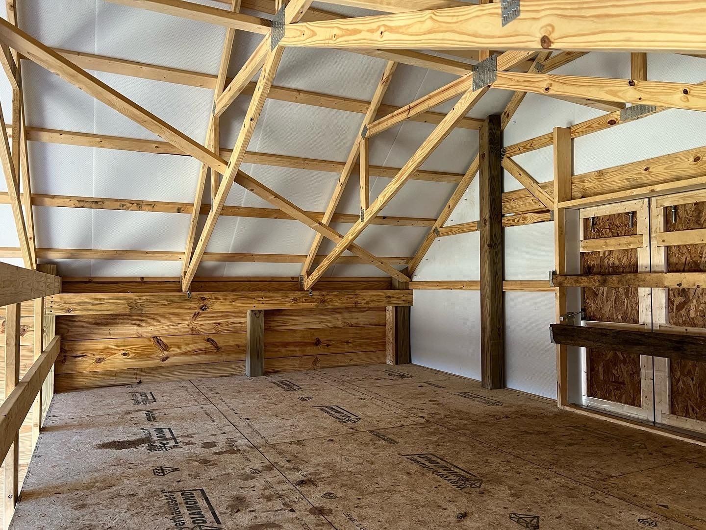 The interior of a wooden loft space featuring exposed beams, rafters, a plywood floor, and light-colored wall panels.
