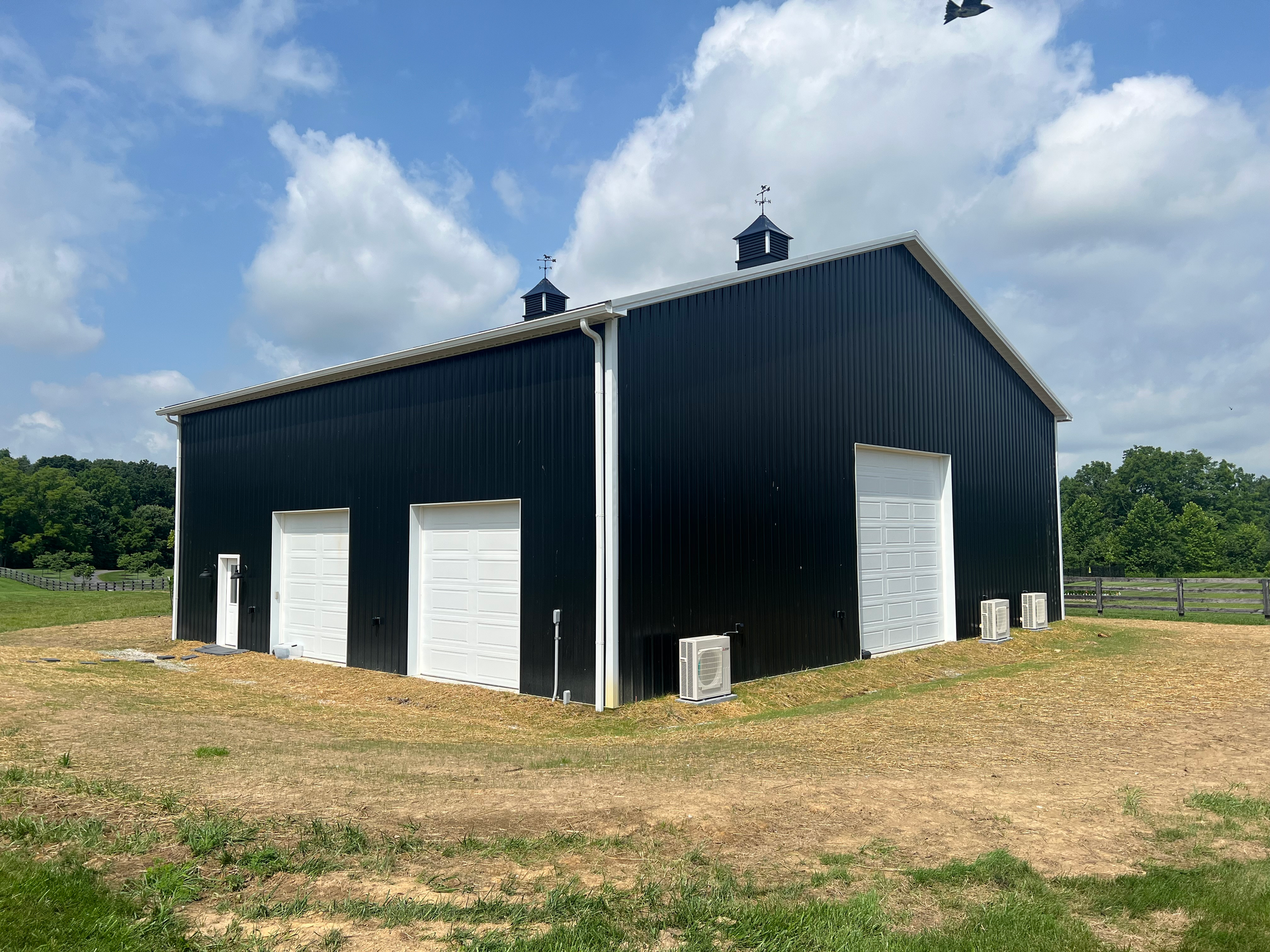 A black, rectangular metal building with white roll-up garage doors and cupolas, set in an open field under a blue sky.