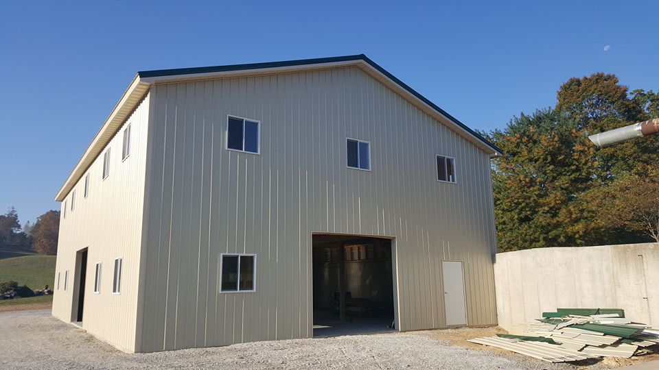 A beige, two-story metal barn with a large open front door, multiple windows, and a black roof in a rural setting.
