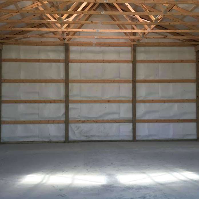 An empty, unfinished pole barn interior with concrete floors, white-faced insulation between studs, and exposed rafters.