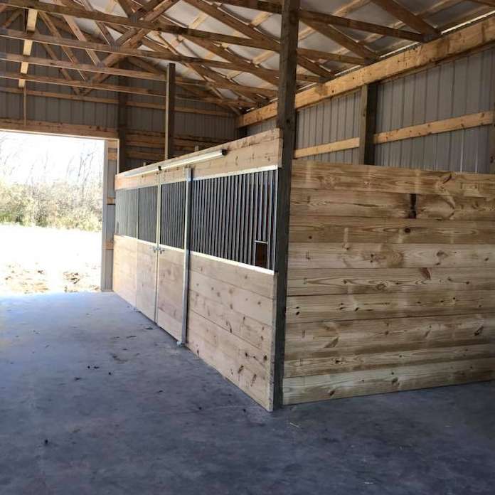 A row of wooden horse stalls with metal barred tops inside a large barn with a concrete floor and open doorway.