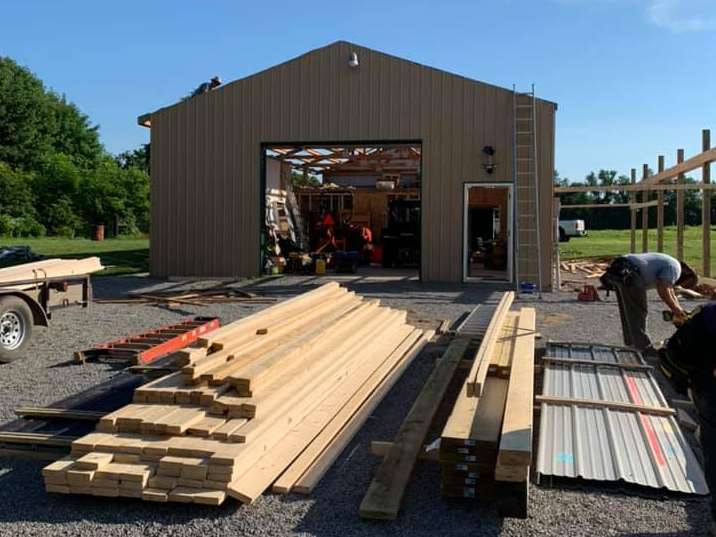 Construction site with a tan building, stacks of lumber, metal roofing, and a worker under a clear blue sky.