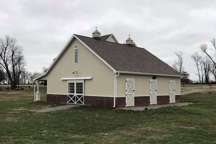 A beige barn with a brown shingle roof, two cupolas, and white trim stands in a grassy field on a cloudy day.