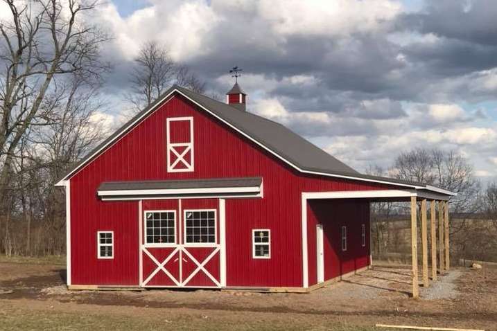 A red barn with white trim, a loft door, and a side open-air shelter on a grassy lot under a cloudy sky.