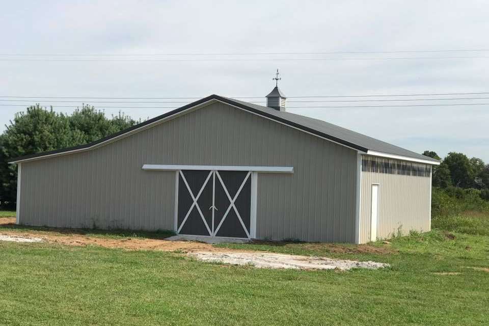 A tan metal barn with a central sliding door, white trim, and a cupola on the roof in a grassy field.