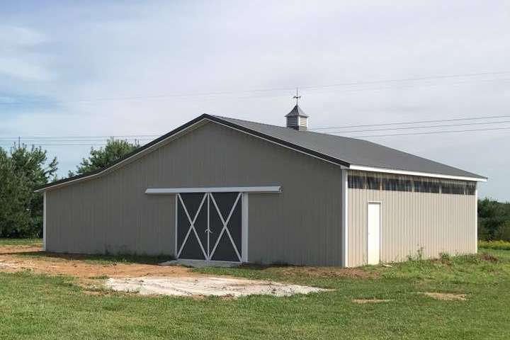 A light gray, single-story barn with a dark metal roof, a sliding cross-buck door, and a white cupola in a grassy field.