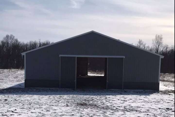 A gray metal pole barn with a central open sliding door stands in a snowy, rural field under a bright sky.