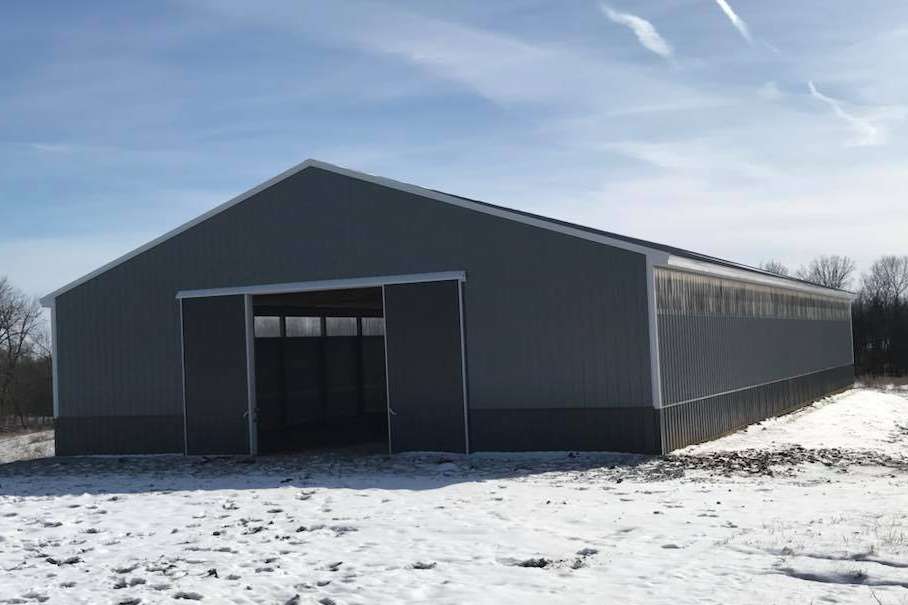 A grey, metal-sided barn with white trim and an open sliding door, situated in a snowy, rural landscape under a blue sky.