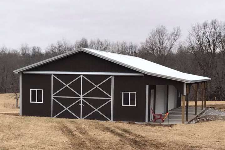 A dark brown pole barn with a white roof, sliding doors, windows, and an open side porch, set in a grassy field.